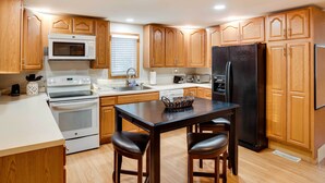A modern kitchen with wooden cabinets, a black refrigerator, a microwave, an oven, and a dining area with two stools.