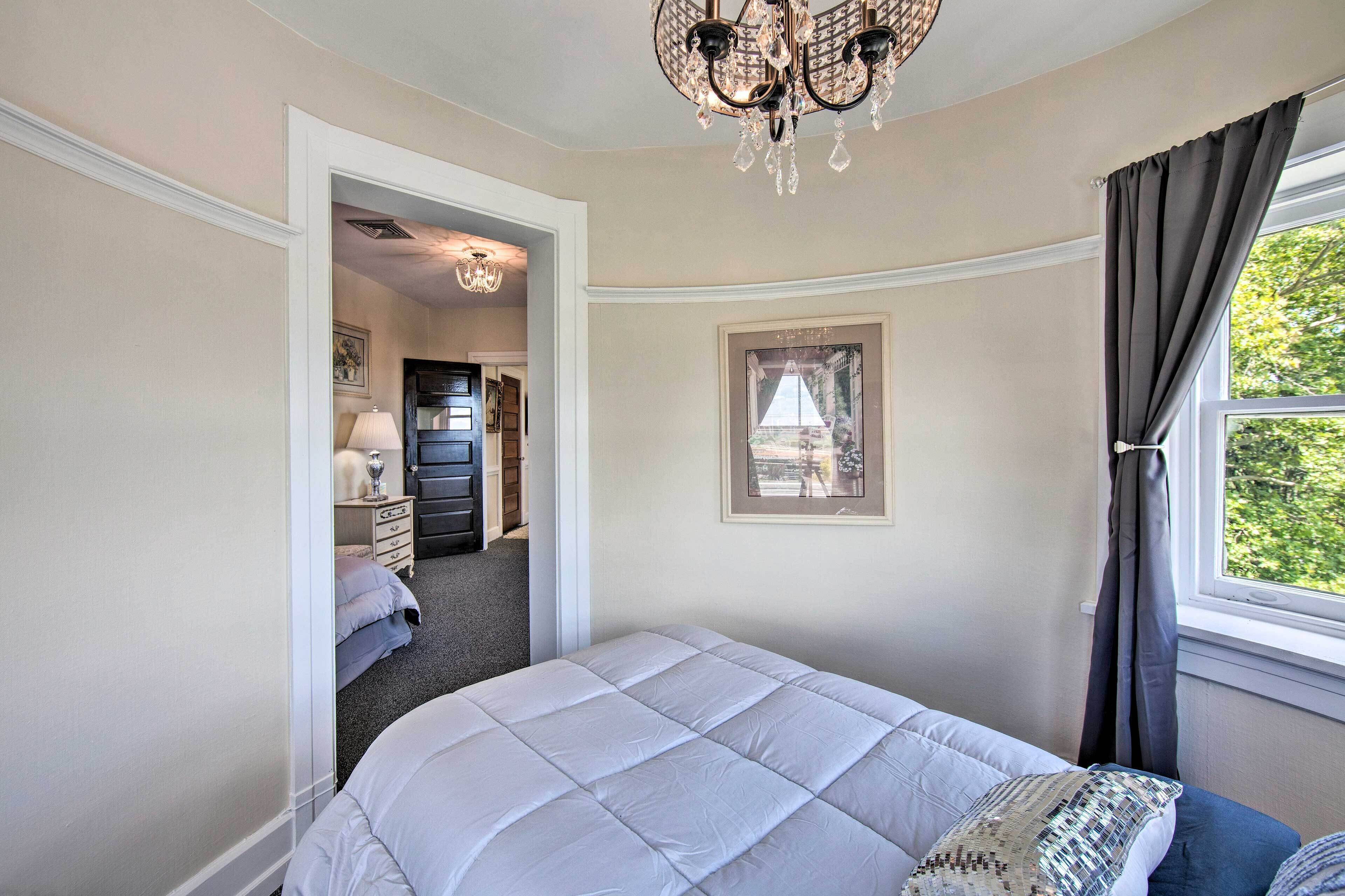 Une chambre à coucher avec un lit, une fenêtre avec des rideaux, un lustre et une porte donnant sur une autre pièce.