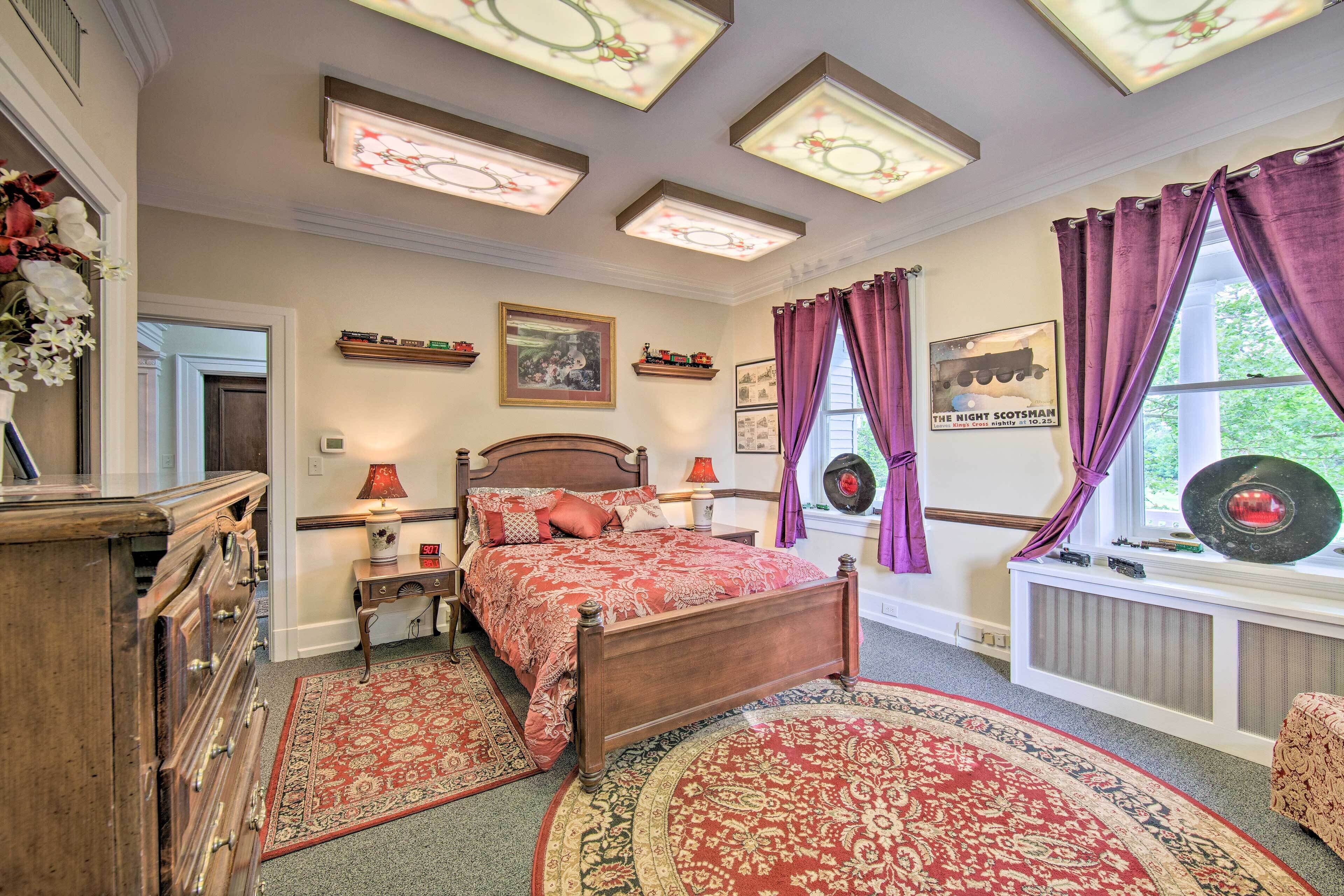 Quarto com cama de madeira, colcha estampada em vermelho e bege, mesa de cabeceira com luminária, cômoda, janela com cortinas roxas e radiador fixado na parede.