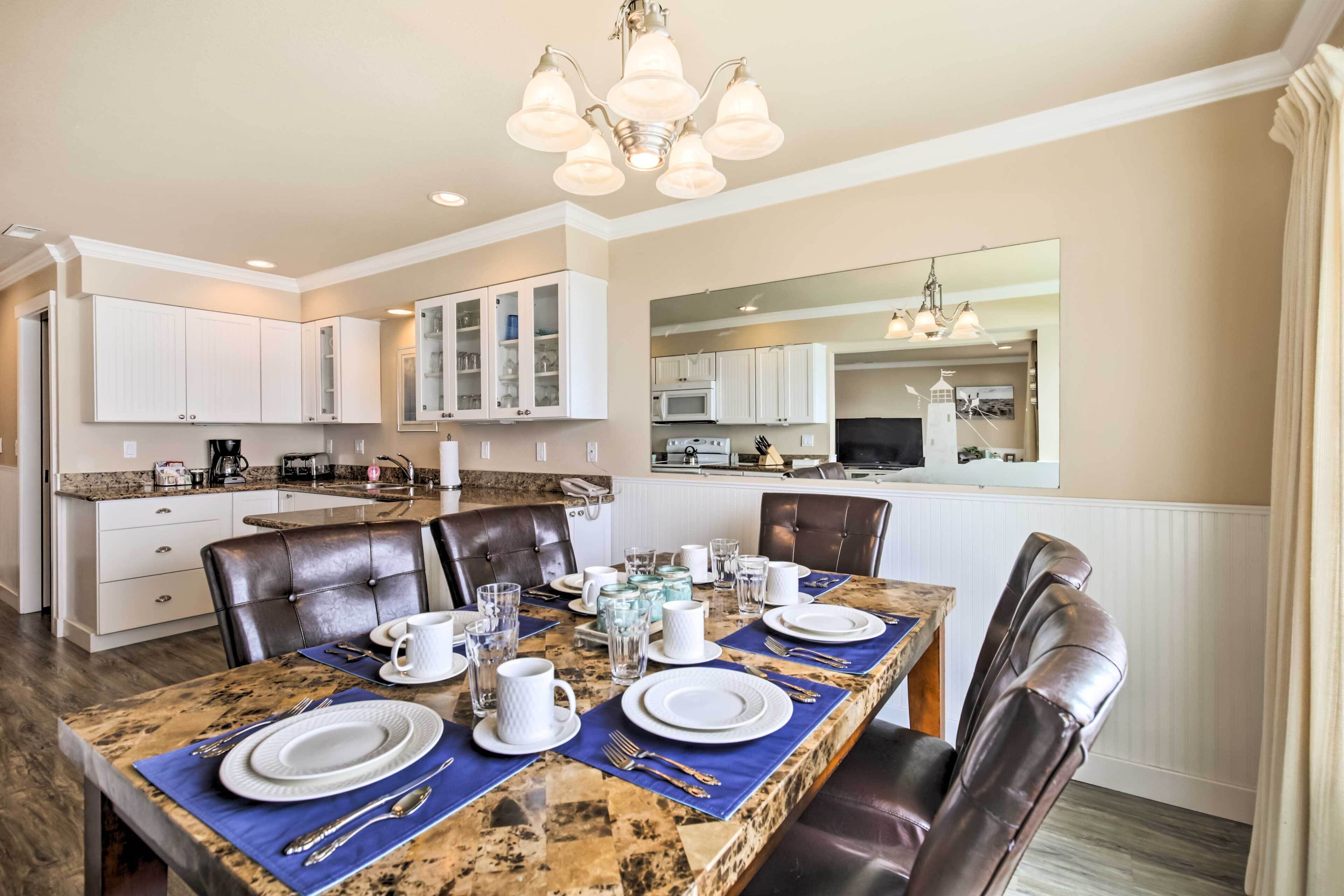 A dining area with a marble table set for four, surrounded by leather chairs. A chandelier hangs above, and the room has white cabinetry and a large mirror on the wall.