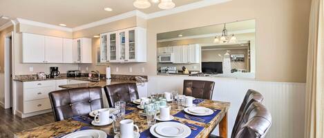 A dining area with a marble table set for four, surrounded by leather chairs. A chandelier hangs above, and the room has white cabinetry and a large mirror on the wall.