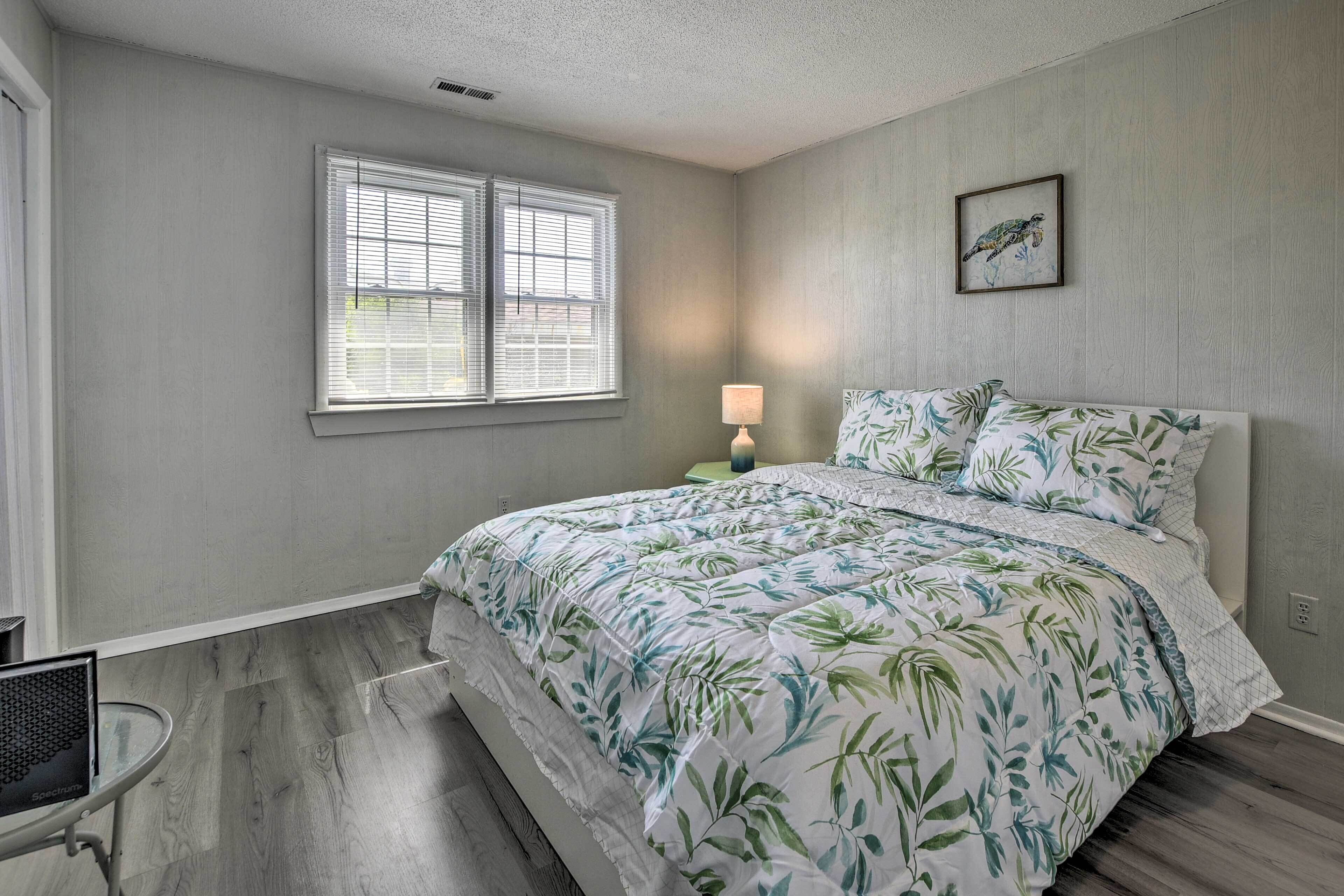 A bedroom with a bed, a window with blinds, a bedside lamp, and a framed picture on the wall.