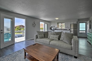 A spacious living room with a grey sofa, wooden coffee table, and patterned cushions. A kitchen with white cabinets and stainless steel appliances is visible in the background.