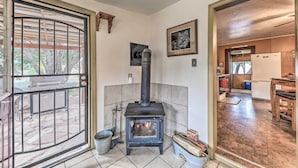 A room with a wood-burning stove, a door leading to an outdoor area, and a kitchen visible through an open door. - Cozy High Rolls Cottage Near Hiking Trails! (High Rolls Mountain Park)