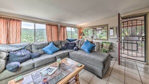 A spacious living room with a grey sectional sofa, a wooden coffee table, and large windows offering a view of greenery. - Cozy High Rolls Cottage Near Hiking Trails! (High Rolls Mountain Park)