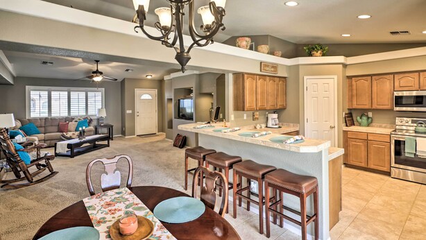 A spacious living room with a large window, a dining area with a wooden table, and a kitchen with wooden cabinets and a central island. - Fort Mohave Home < 8 Mi to Colorado River! (Fort Mohave)