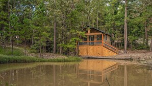 Une cabane en bois dotée d’un balcon, entourée de forêt et bordée d’un étang.