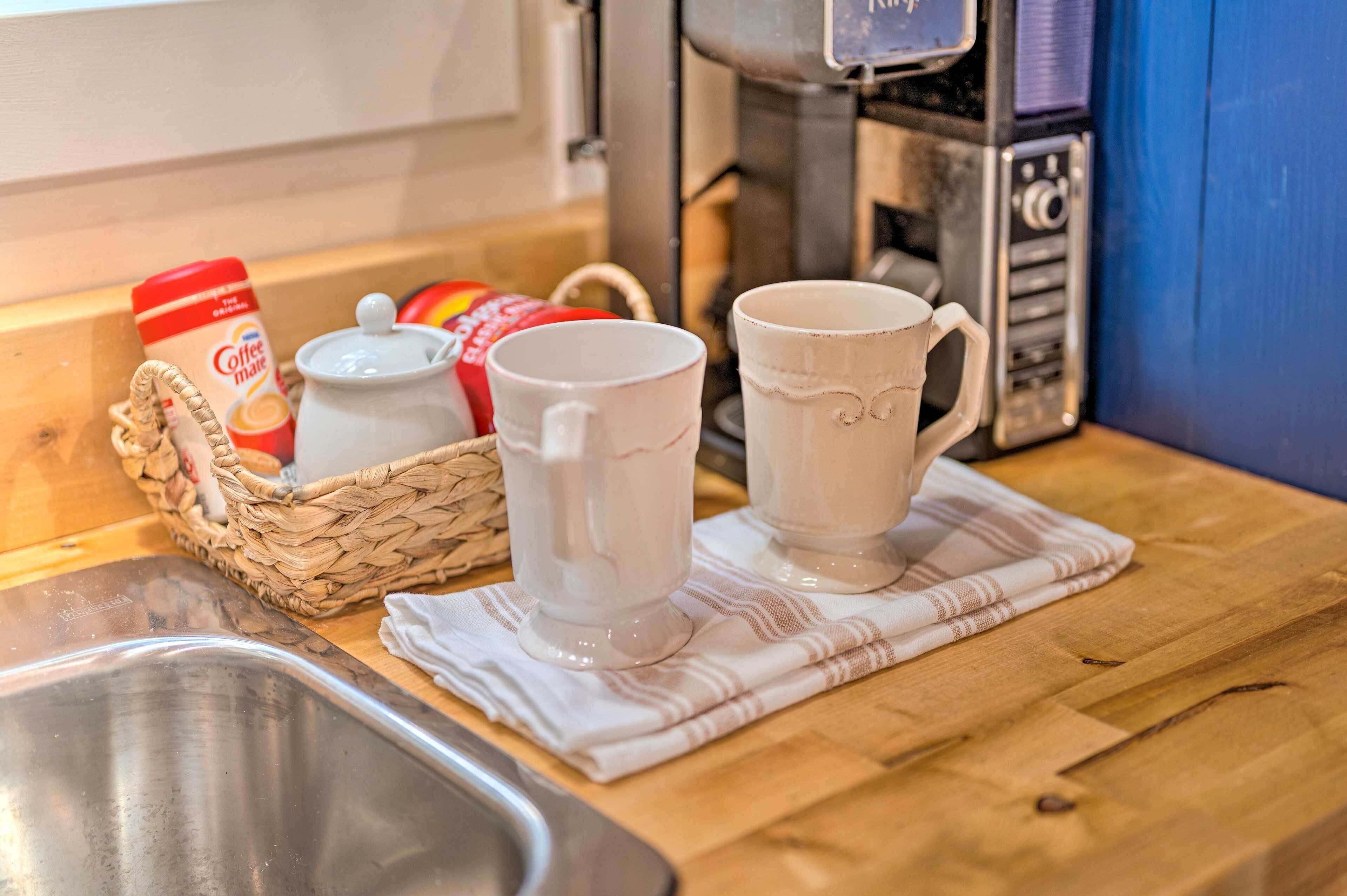A kitchen counter with two white mugs, a coffee maker, and a basket containing a coffee can and a jar.