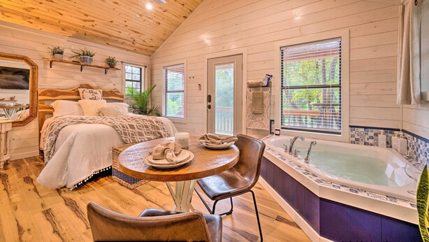 L’intérieur chaleureux d’un chalet avec un plafond en bois, un lit recouvert d’une couverture à motifs, une table ronde en bois entourée de chaises et une grande baignoire dont le pourtour est recouvert de carreaux.