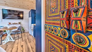 Une pièce avec un plafond en bois, un coin repas avec une table et des chaises, un coin cuisine avec des placards et une fenêtre, et un mur coloré orné de motifs géométriques.