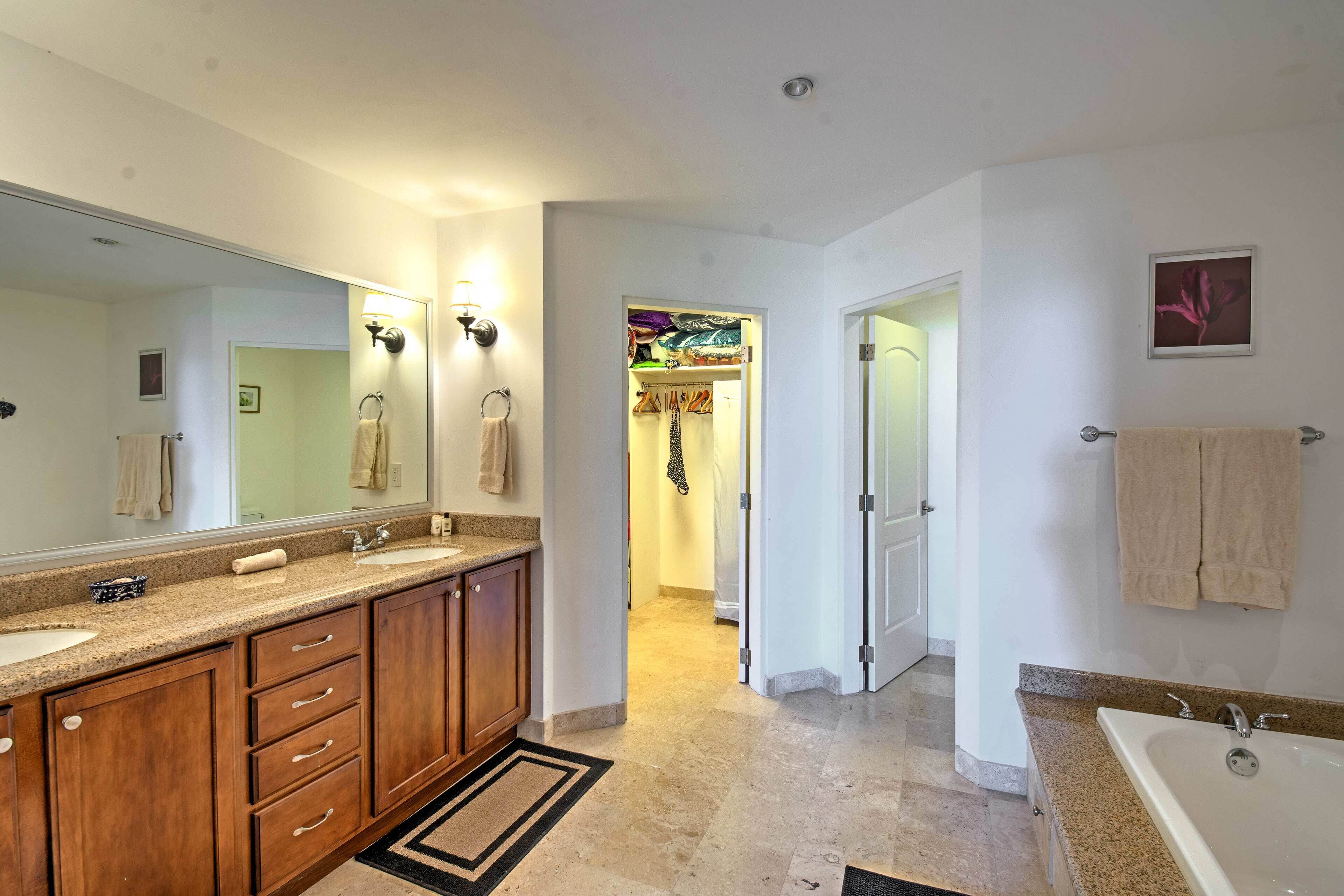 A modern bathroom with a double sink, vanity, and a large mirror. There is a walk-in closet with hanging clothes and a door leading to another room.
