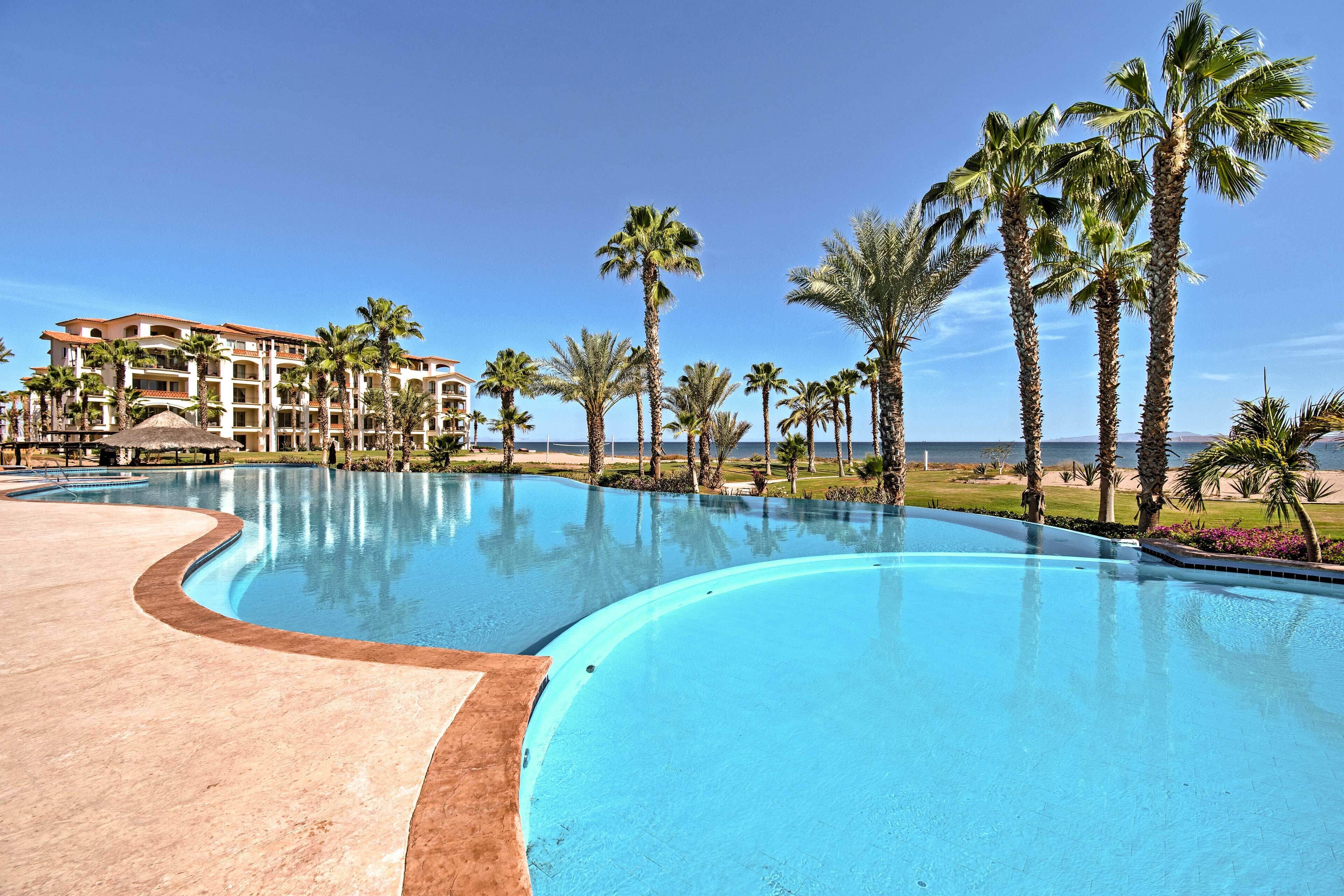 A resort pool with a curved edge, surrounded by palm trees and a building with multiple balconies.