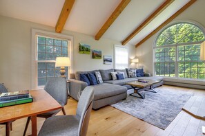 Un salon comprenant un canapé, des fauteuils, une table basse et une table à manger. Il y a deux fenêtres, l’une donnant sur des arbres et l’autre sur des montagnes.