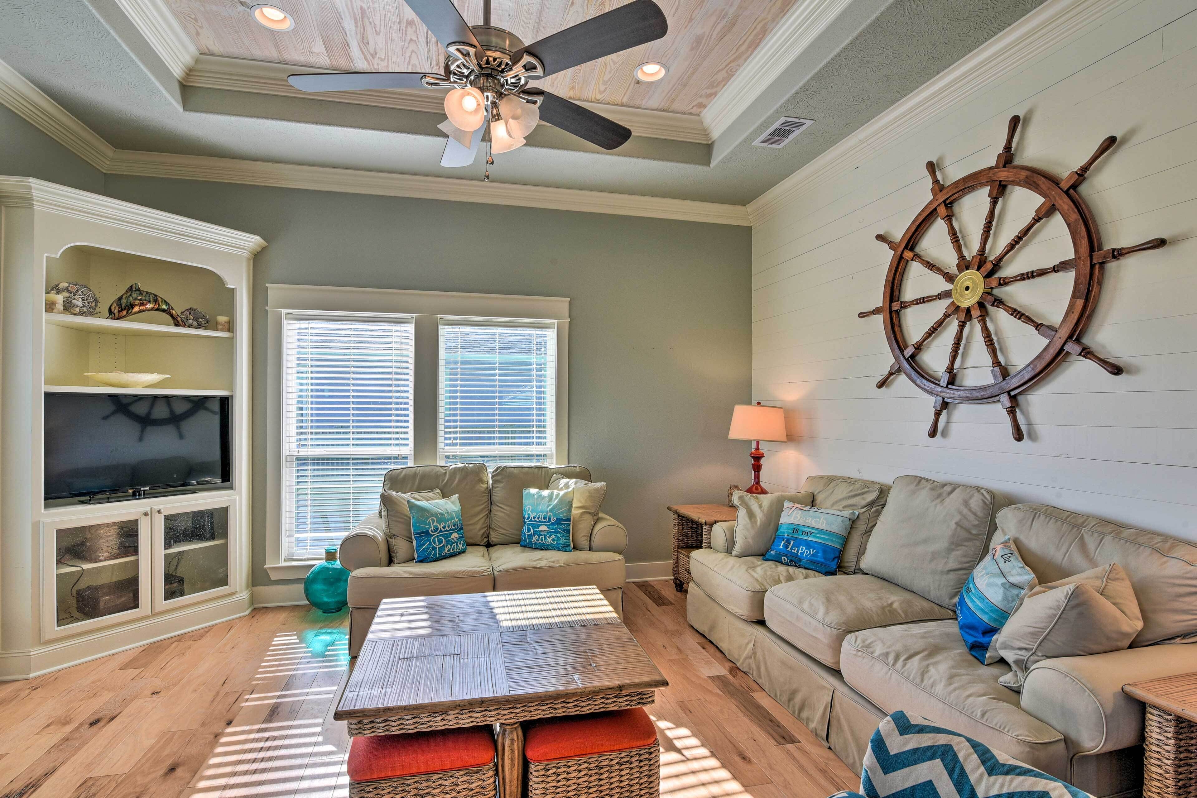 Ein Wohnzimmer mit Deckenventilator, einer großen hölzernen Schiffsschrauben-Dekoration, einem beige gefärbten Sofa mit blauen Kissen, einem Fernseher auf einer Kommode und einem Tisch mit einer Tischläufer mit Streifen.