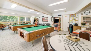A pool table, a billiards table, and a chess table in a well-lit room with windows and a clock on the wall.