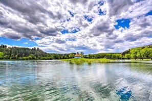 A lake with a house on a hill, surrounded by trees and a cloudy sky. - Charming Rapid City Apartment: Walk to Lake! (Rapid City)