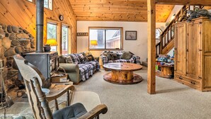 A cozy cabin interior with a stone wall, a wood-burning stove, a plaid sofa, a round wooden table, and a staircase leading to a loft. - Truckee Winter Escape 6 Mi to Ski Slopes! (Truckee)