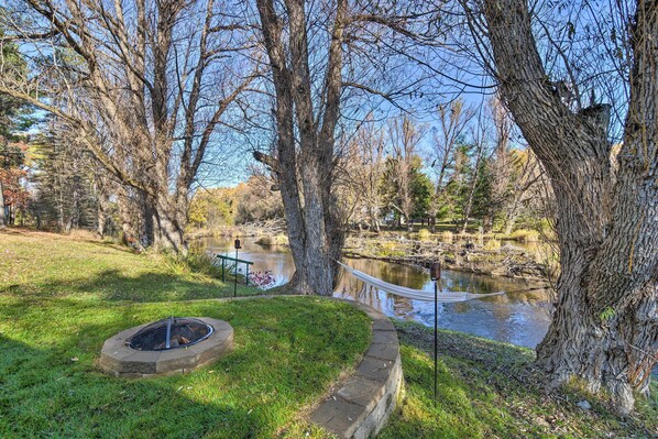 A serene outdoor area with a fire pit, a small pond, and a hammock. - Riverfront Grayling Retreat w/ Fire Pit & Dock! (Grayling)