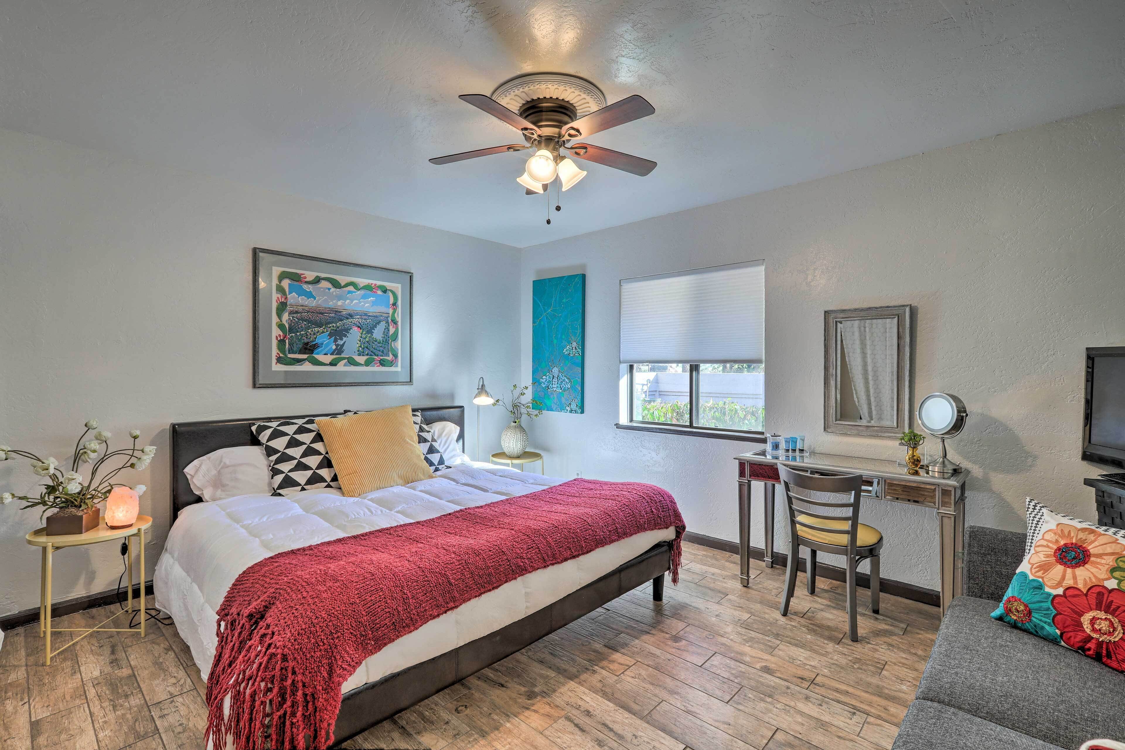 A bedroom with a bed, a desk, a chair, a ceiling fan, a window, and a TV.