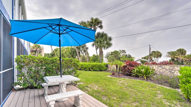 A patio with a white table and bench, a blue umbrella, and a garden with various plants and a stone wall. - Longboat Key Condo W/lanai, Walk to Beach & Shops (Longboat Key)