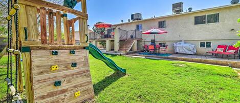 Un patio trasero con una estructura de madera para escalar, un tobogán y una zona de piscina con sillas y sombrillas rojas.