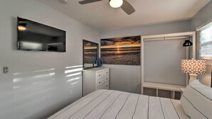A bedroom with a bed, a ceiling fan, a TV mounted on the wall, a nightstand, a lamp, and a painting of a sunset. - Charming Vacation Rental: Close to Downtown! (Sarasota)
