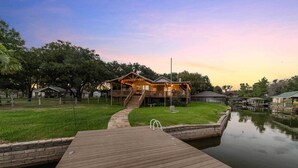 Une maison en bois dotée d’une terrasse et d’un quai s’avançant dans une étendue d’eau calme, le tout entouré de verdure et d’autres bâtiments.
