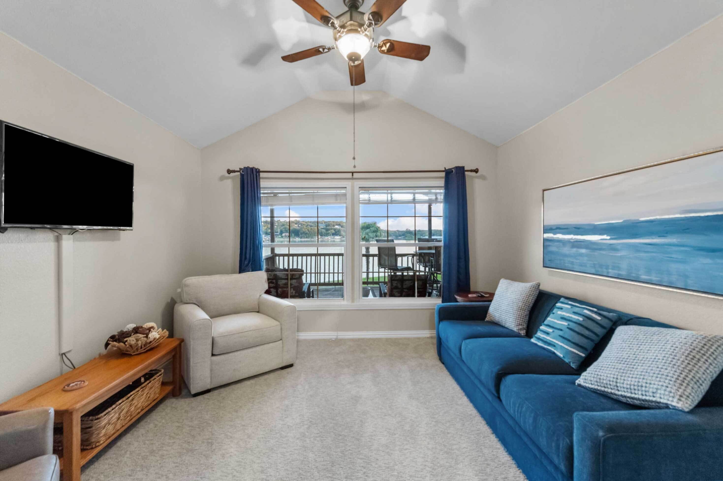 A living room with a ceiling fan, a flat-screen TV, a sofa, a blue couch, and a large window with curtains.