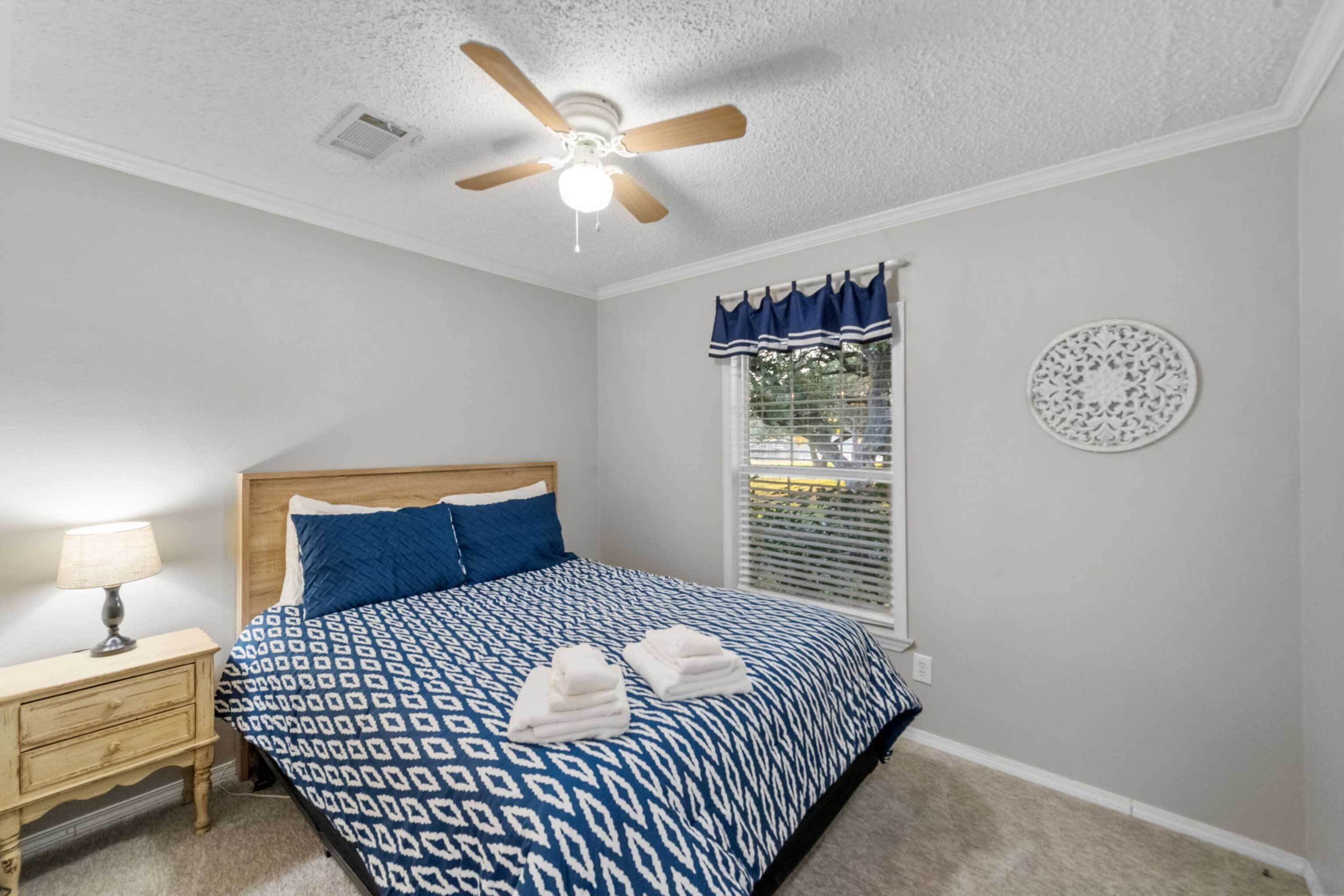 Ein Schlafzimmer mit Bett, Nachttisch, Deckenventilator, Fenster mit Jalousien und einer Wanddekoration.