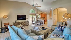 A spacious living room with a large sofa, a coffee table, and a TV. There is a dining area with chairs and a table, and a kitchen in the background.