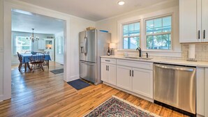 A modern kitchen with stainless steel appliances, white cabinetry, and a dining area with a wooden table and chairs. - Historic Family Home < 1 Mi to Long Beach! (Oak Island)