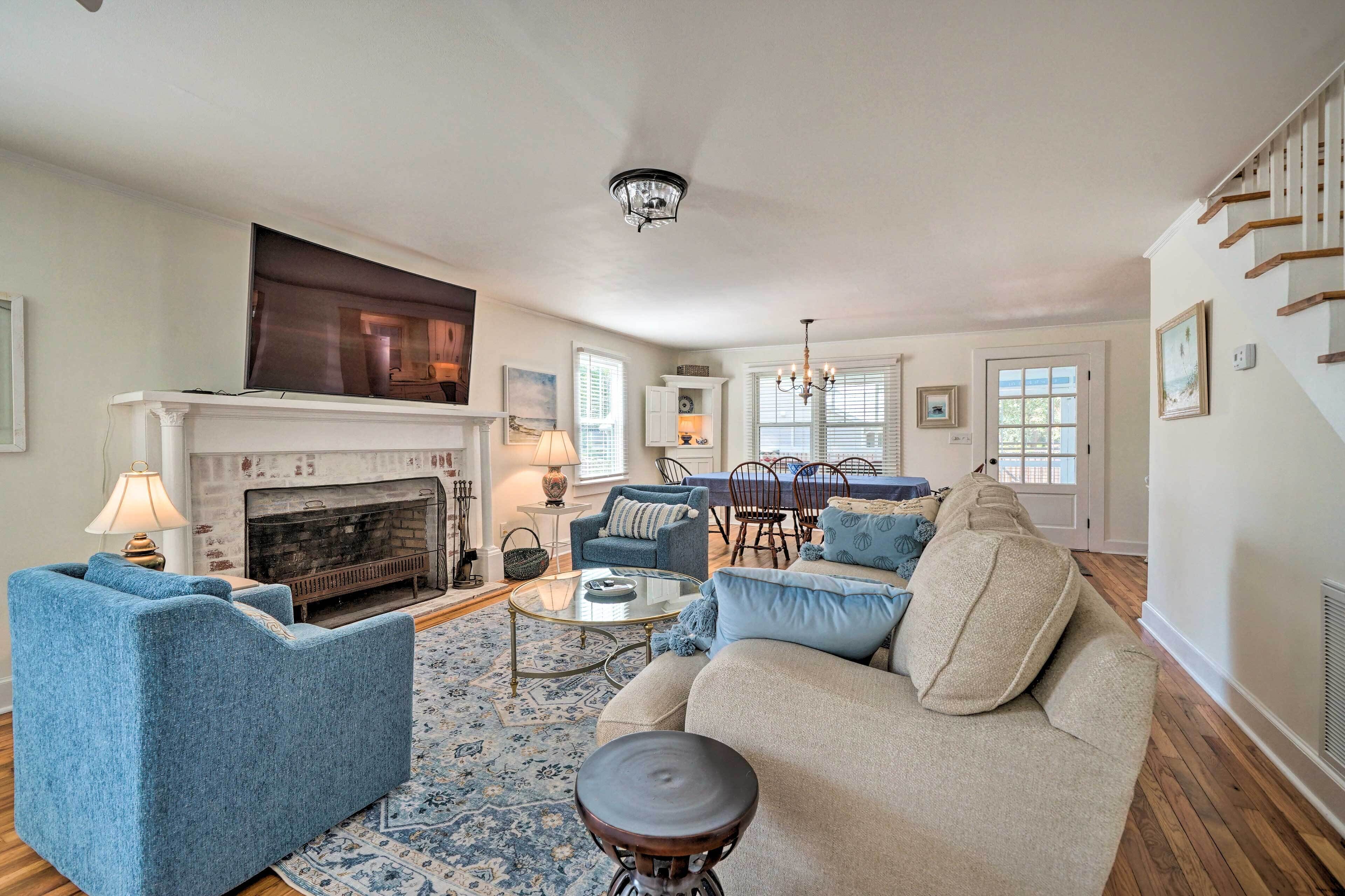 A living room with a fireplace, blue and beige furniture, a dining area with a table and chairs, and a staircase.