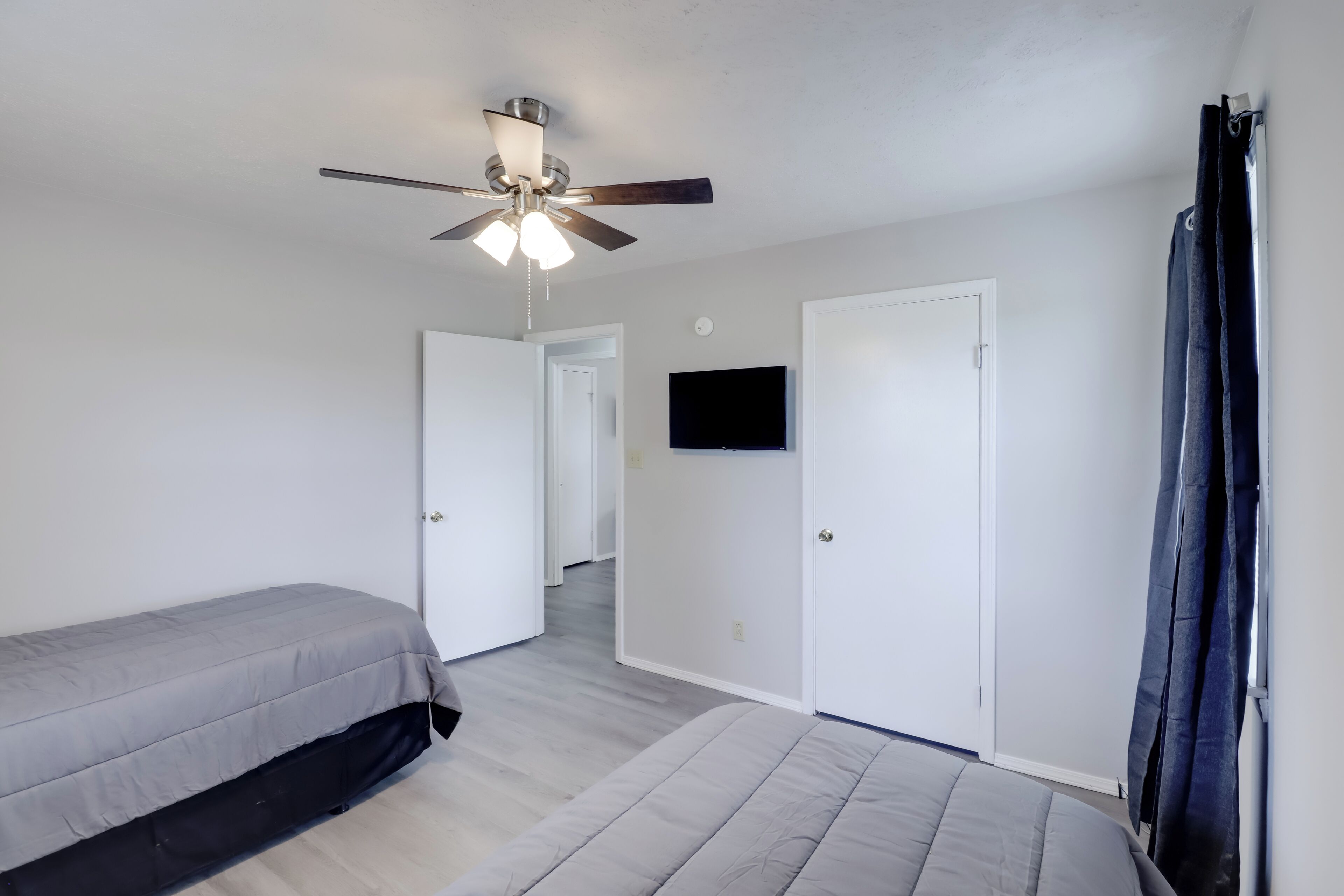 A bedroom with two beds, a ceiling fan, a TV mounted on the wall, and a door leading to another room.