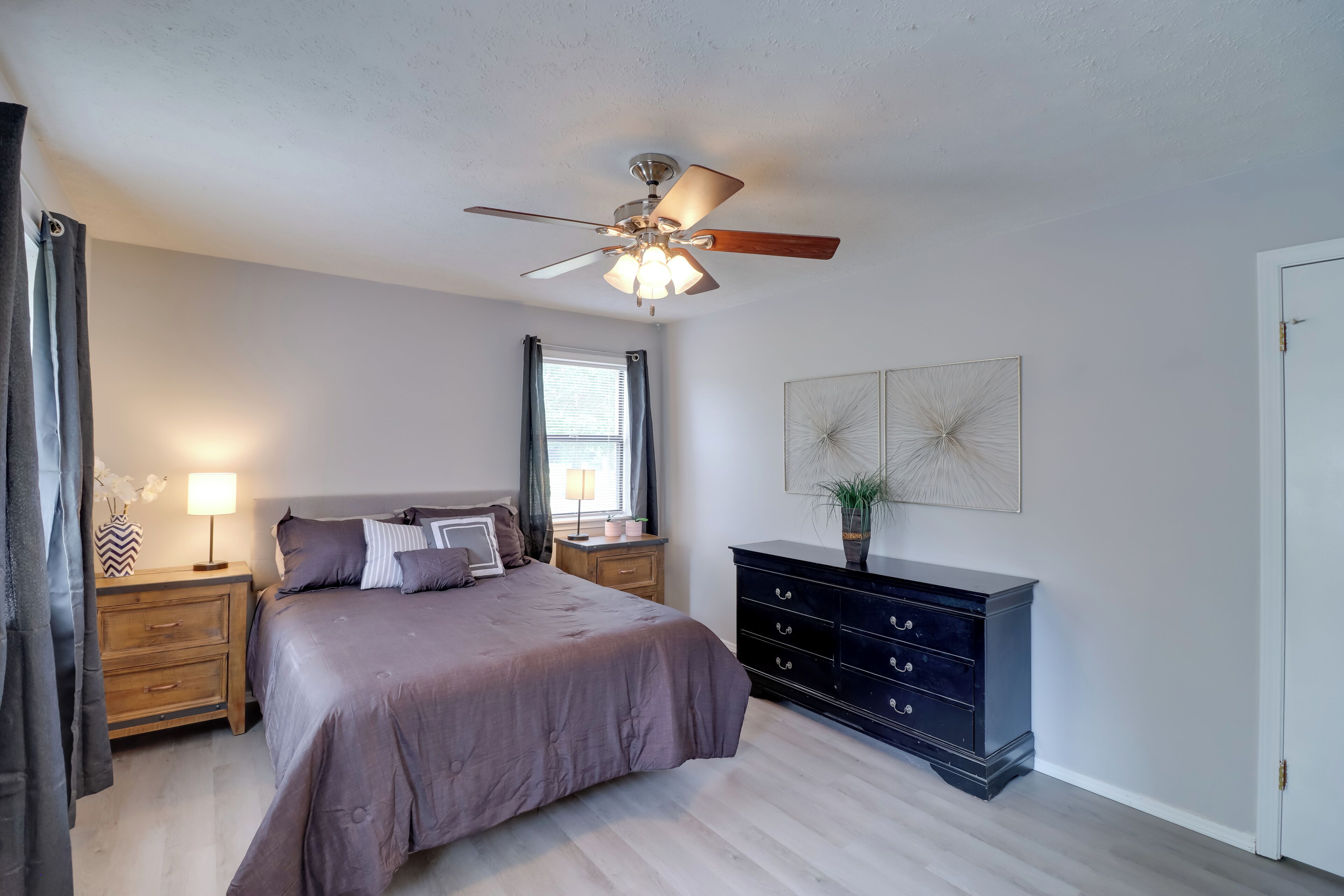 A bedroom with a bed, nightstands, a dresser, a ceiling fan, and a window with curtains.