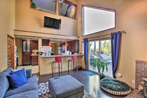 A modern living room with a grey sofa, ottoman, and a pet bed. A kitchen with a bar area is visible in the background. - Puget Sound Home w/ Beautiful Yard & Deck! (Des Moines)