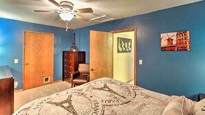Ein Schlafzimmer mit einem Bett mit Paris-Motiv, einem Deckenventilator, einer Kommode, einem Stuhl und einer Tür zu einem anderen Raum.