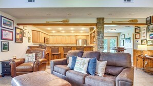 A spacious living room with a brown leather sofa, a matching ottoman, and a wooden coffee table. There is a kitchen area visible through an open doorway, featuring wooden cabinets and a stainless steel refrigerator. - 'troutfest' Riverfront Norfork Home, Great Fishing (Norfork)