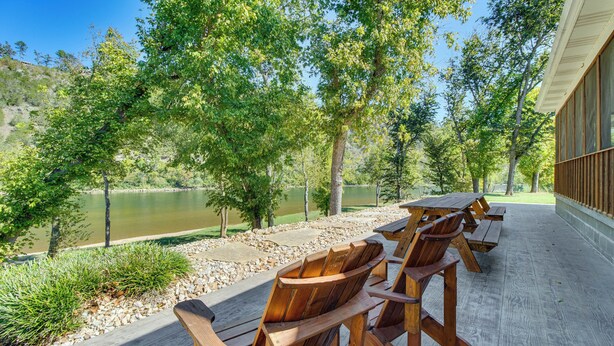 A wooden deck with chairs and tables overlooking a lake, surrounded by trees and a building with a wooden facade. - 'troutfest' Riverfront Norfork Home, Great Fishing (Norfork)