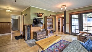 A spacious living room with a wooden coffee table, a television mounted on the wall, a bookshelf, and a large window with a view of the outdoors. - Remote Escape: Riverside Home w/ Scenic Views (Idanha)
