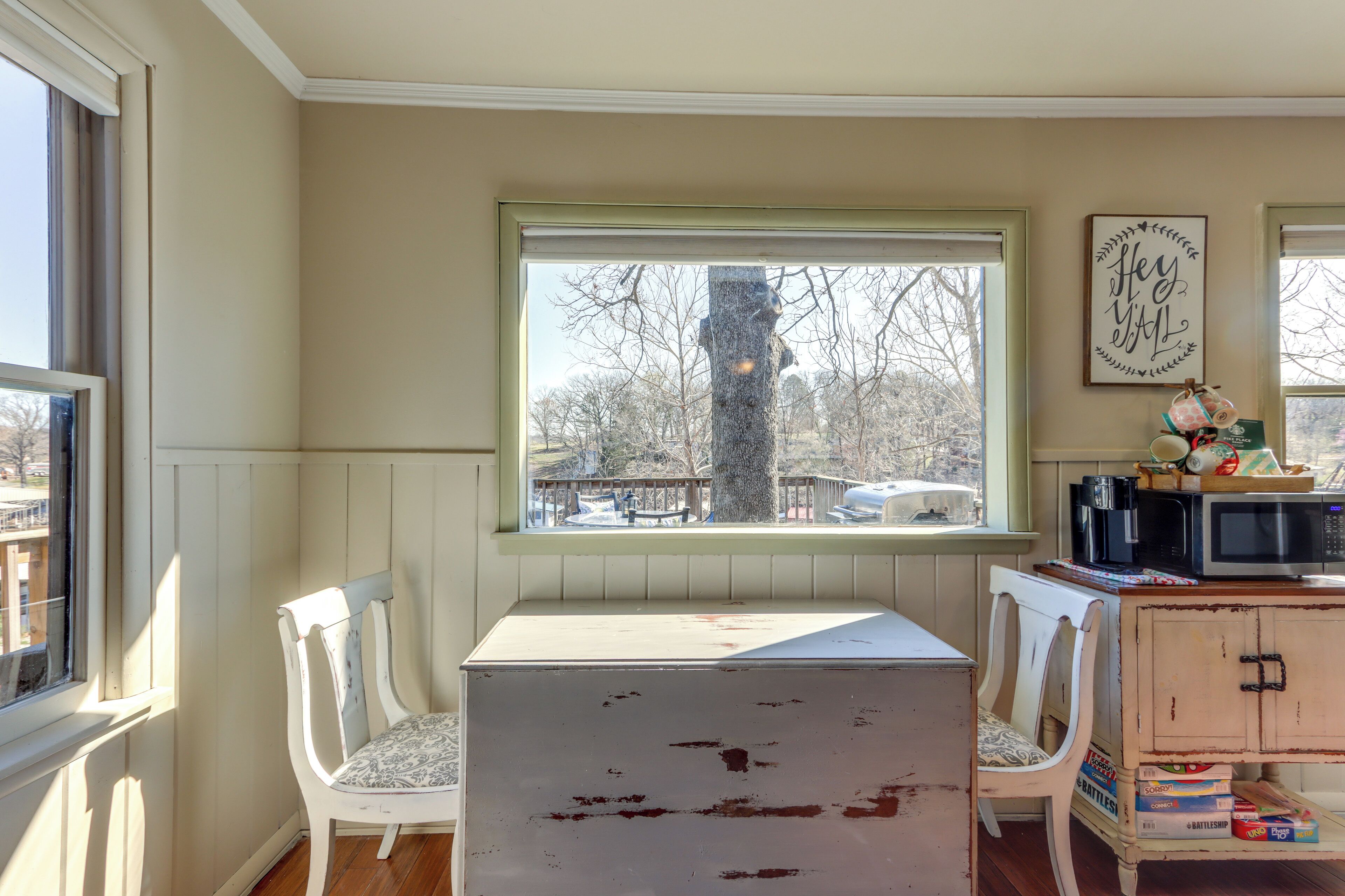 Una habitación con ventana, dos sillas blancas, una mesa, un mueble con electrodomésticos de cocina y un cartel en la pared.