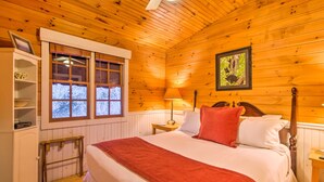 A wooden-paneled bedroom with a bed, a nightstand, a lamp, a shelf with decorative items, and a window with a view of trees. - Quaint Bryson City Cottage w/ Smoky Mountain Views (Bryson City)