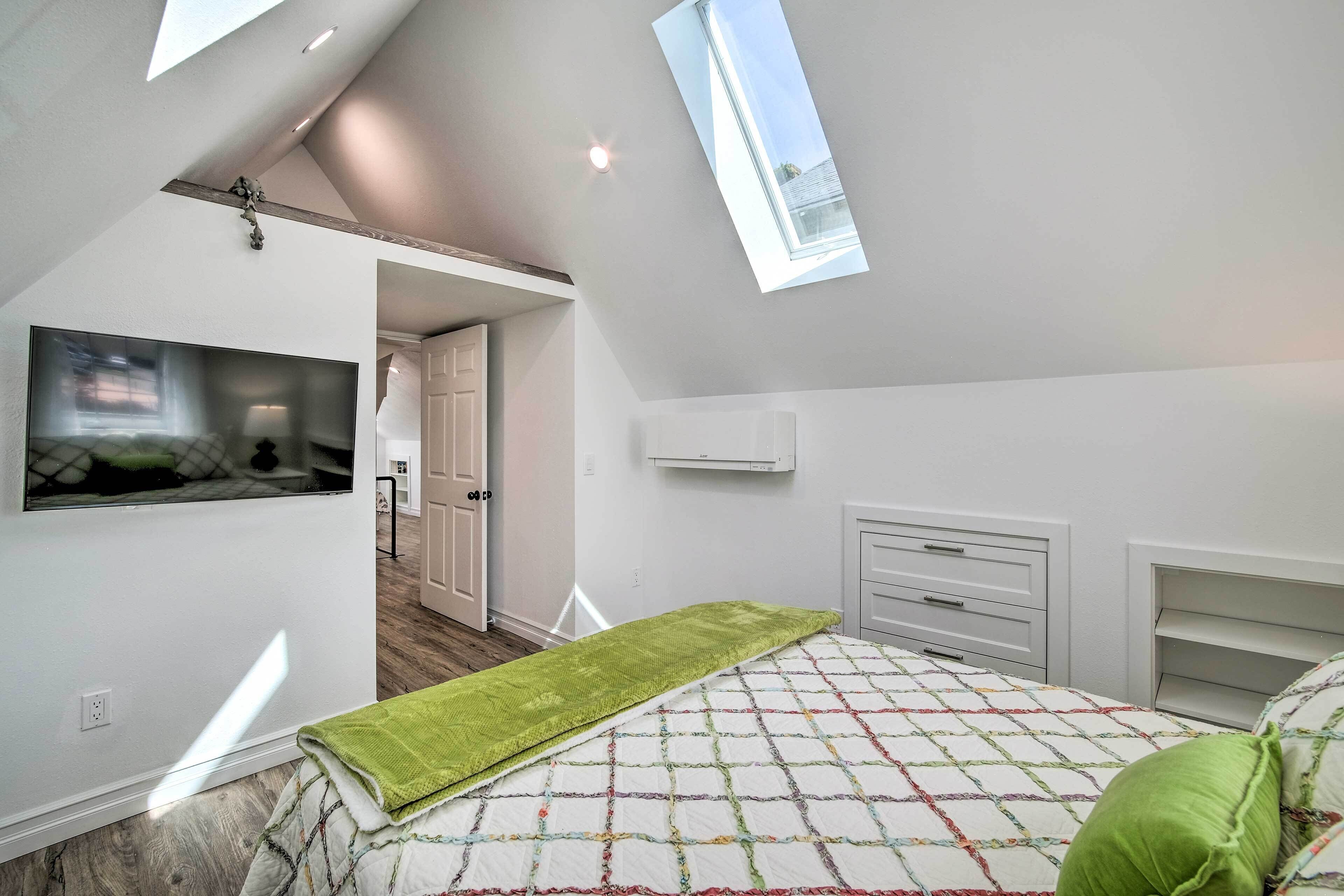 A bedroom with a sloped ceiling, skylight, a bed with a quilt, a green throw, a TV, and a white dresser.
