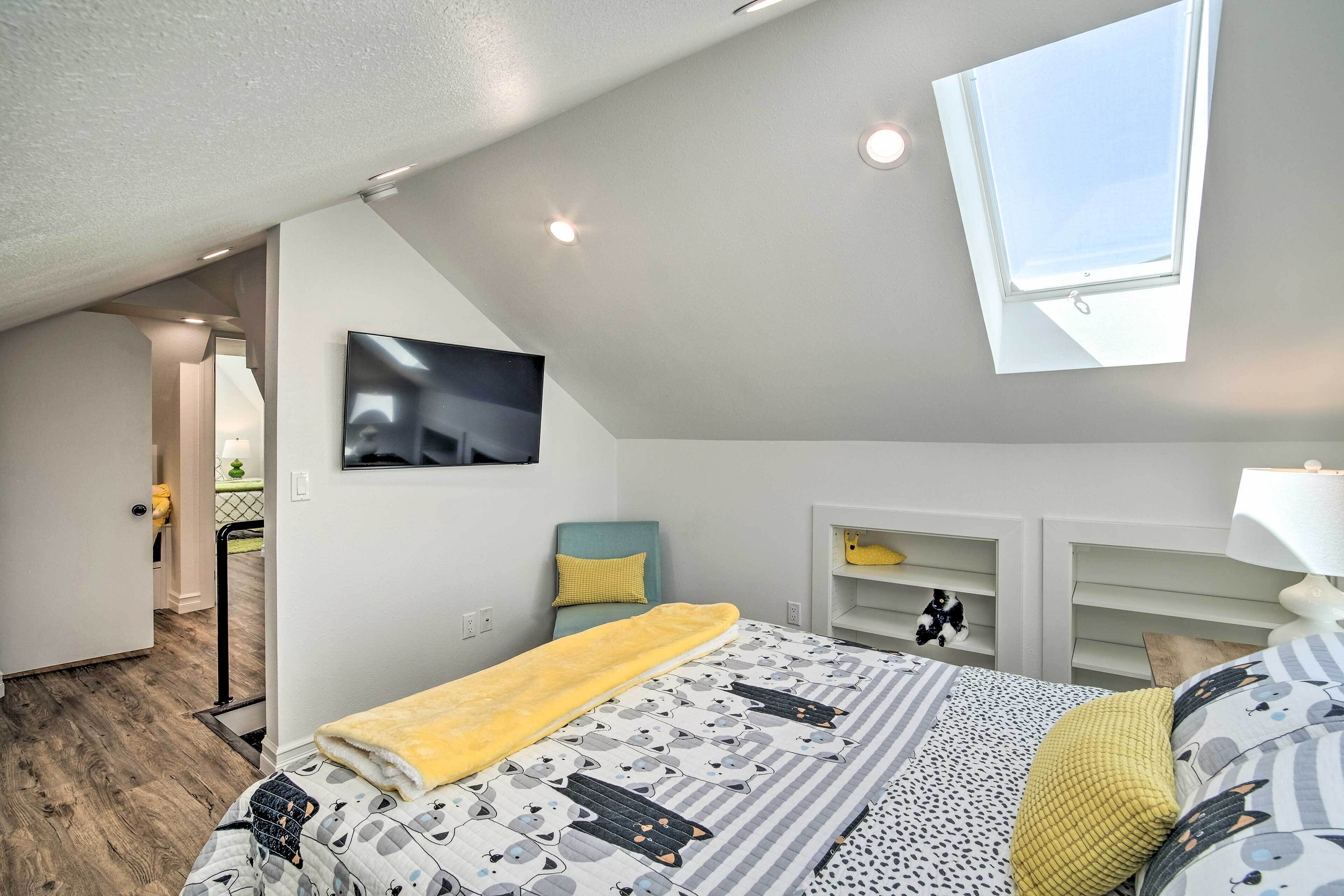 A bedroom with a sloped ceiling, a bed with a patterned bedspread, a yellow blanket, a TV mounted on the wall, and a skylight.