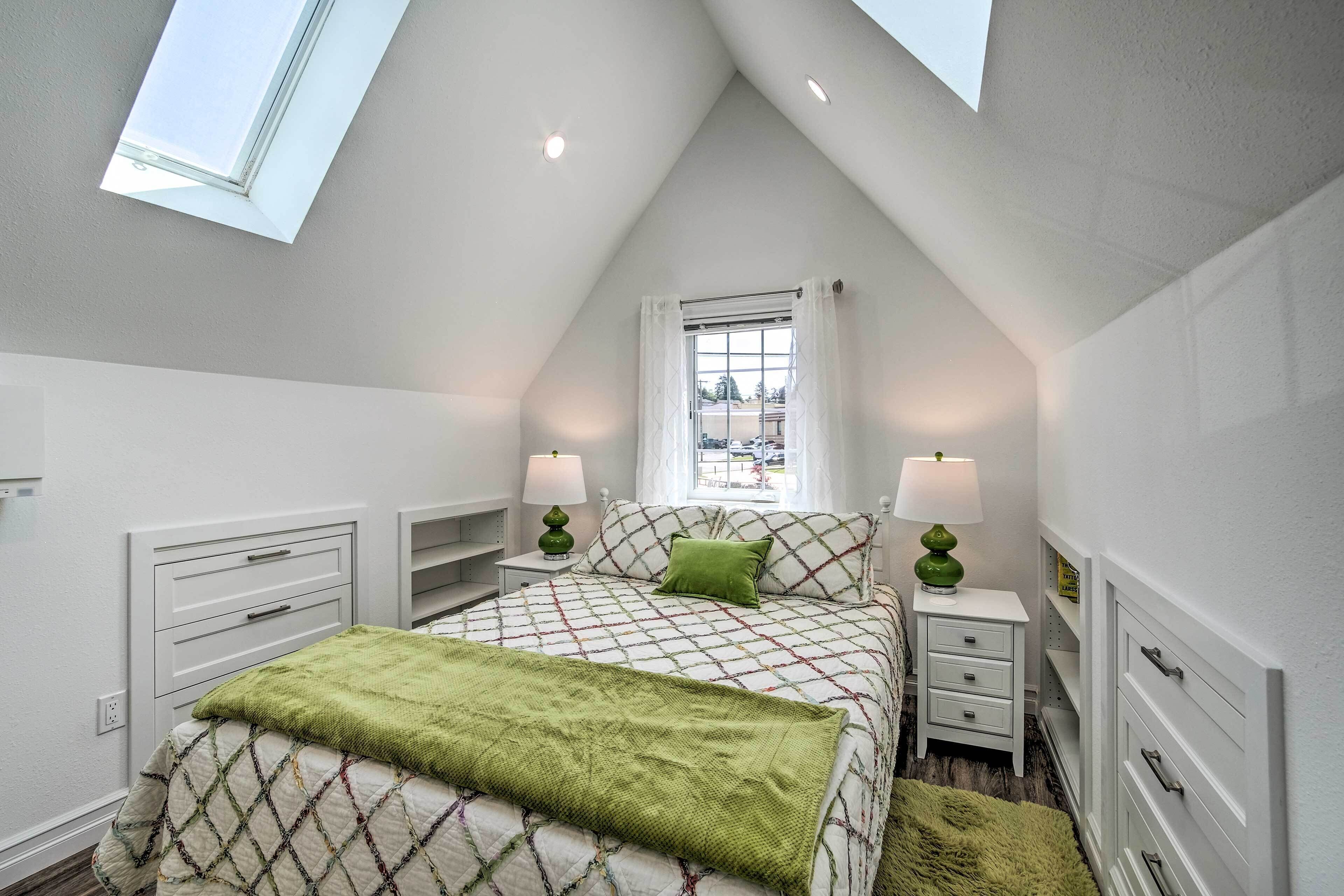 Une chambre avec un plafond en pente, un lit recouvert d’une couvre-lit à motifs, deux tables de chevet avec des lampes, une fenêtre avec des rideaux et une vue sur l’extérieur.