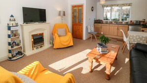 Ein Wohnzimmer mit Kamin, Fernseher, einer Couch, einem Couchtisch und einer Küchenzeile mit Holzausbau und einem Fenster.