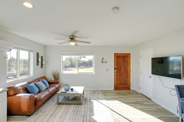 A spacious living room with a brown leather sofa, a coffee table, a ceiling fan, and a large TV. - Peaceful Peoria Bungalow w/ Grill & Pool! (Peoria)
