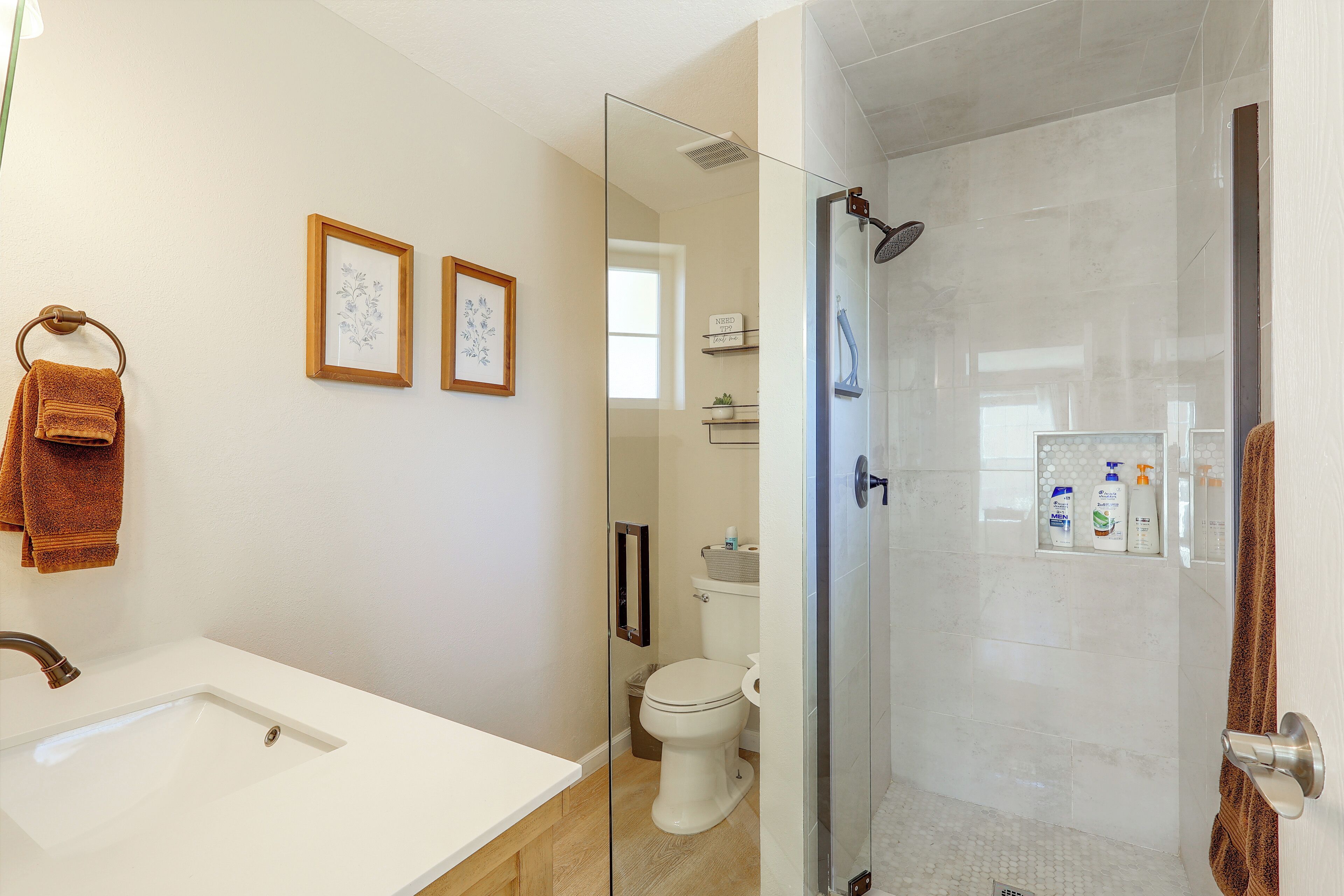 A bathroom with a glass shower enclosure, a white sink, and a toilet. There are framed pictures on the wall and a towel rack with a brown towel.