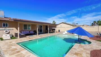 Une piscine avec une terrasse carrelée, un patio couvert aménagé avec du mobilier d’extérieur et un ciel d’un bleu limpide.