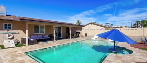 A swimming pool with a tiled deck, a covered patio area with outdoor furniture, and a clear blue sky.