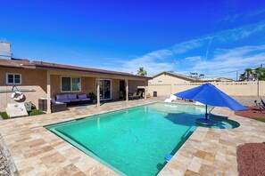 A swimming pool with a tiled deck, a covered patio area with outdoor furniture, and a clear blue sky.